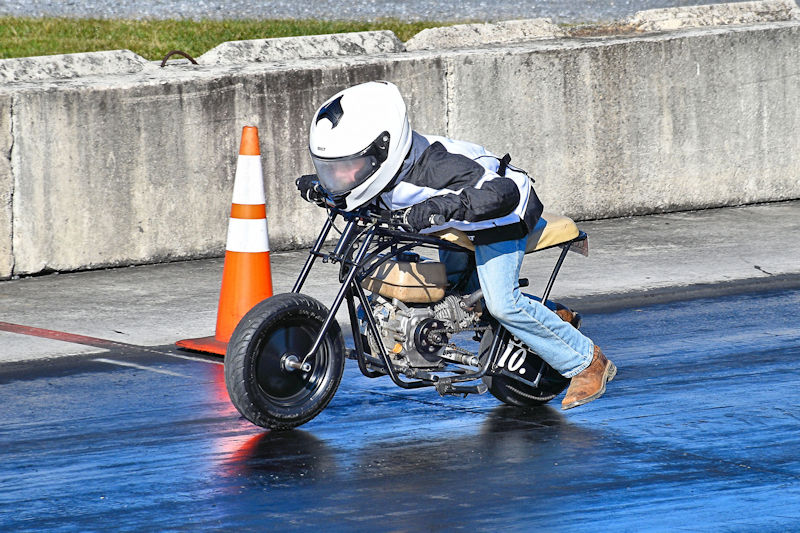 A child racing on a small motorcycle in a protective helmet and gear, leaning forward for balance. A traffic cone is visible in the background on a racing track.