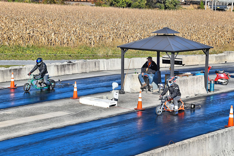 Two children riding miniature motorcycles on a racetrack, with safety cones marking the lanes. A man is sitting under a gazebo observing the racers. Fields of corn are visible in the background.