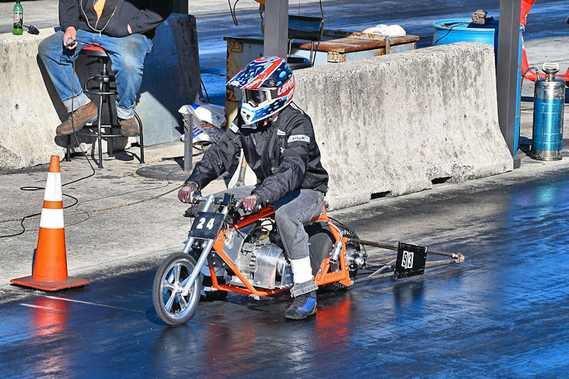 A person wearing a helmet and black jacket is riding a small motorcycle on a drag racing track, with a number '24' displayed on the vehicle. A bystander is seated nearby, and traffic cones are placed along the side of the track.