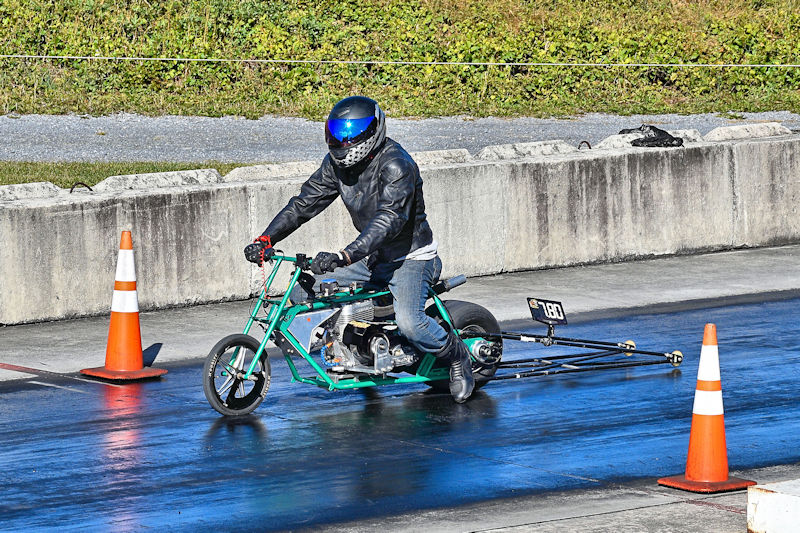 A person riding a small, custom motorcycle on a drag racing track, wearing a helmet and leather jacket, with orange cones marking the track boundaries.