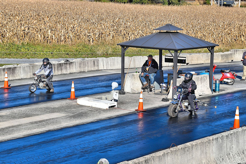 Two children racing mini motorcycles on a drag strip, with orange traffic cones marking the lanes and a gazebo sheltering an observer in the background. A cornfield is visible beyond the track.