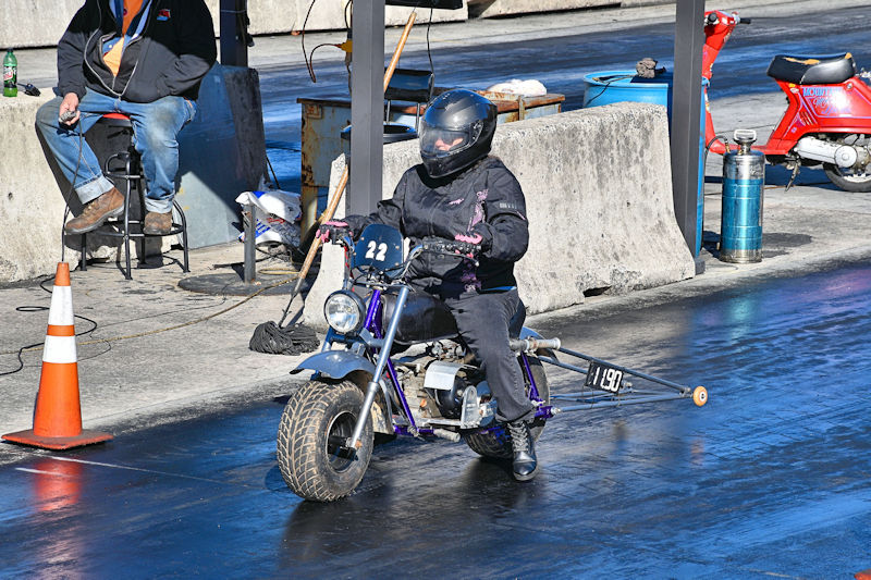 A person in black riding a small motorcycle on a drag strip, wearing a helmet and gloves, with the number 22 displayed on the bike.