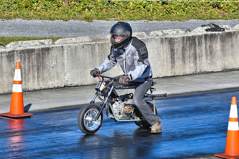 A person in a helmet and protective jacket riding a small motorcycle on a drag racing track, with orange traffic cones lining the course.
