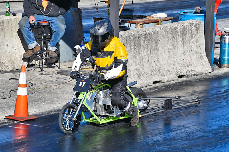 A drag racer in a full-face helmet and yellow and black racing suit rides a small motorcycle during a race, with a spectator sitting nearby and track barriers visible in the background.