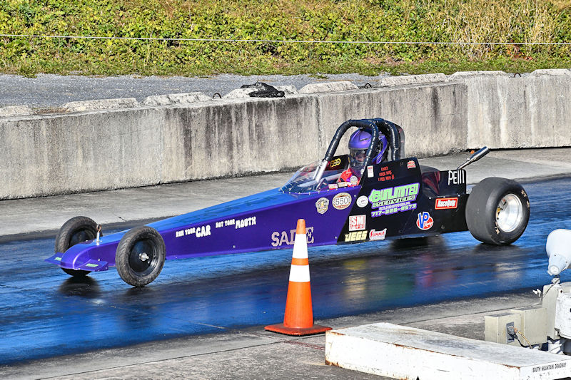 A drag racing car with a purple body and various sponsor decals, positioned on a track with a traffic cone in the foreground.