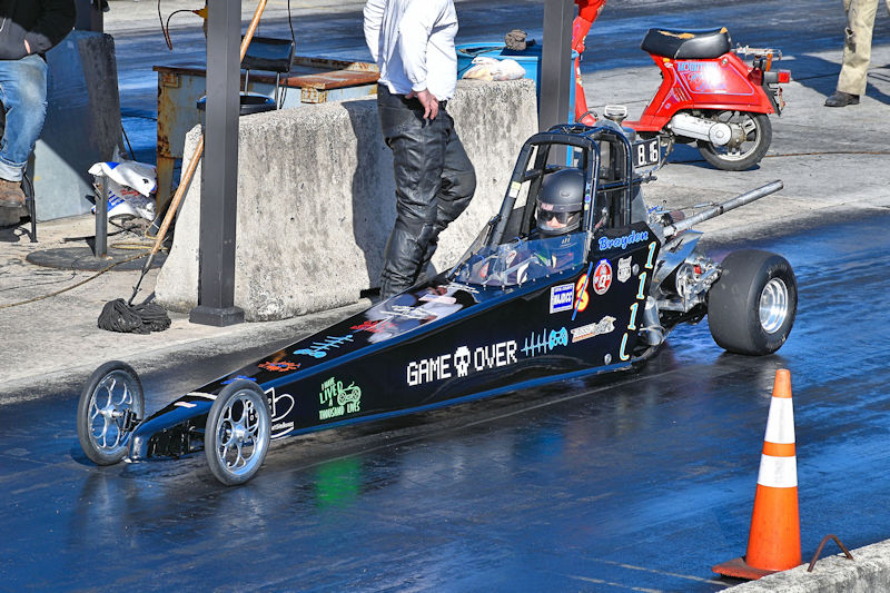 A small black drag racing car with the words 'GAME OVER' on its side, standing on a racetrack. A driver wearing a helmet is seated inside, while a person in black leather pants is beside the car. In the background, there is a small scooter and other people preparing for racing.
