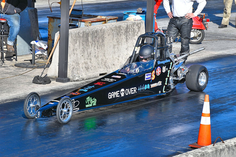 A black drag racing car with various decals, including 'GAME OVER', is positioned on a track, ready for a run. The driver is wearing a helmet and focused on the race. Nearby, a person stands watching, while another is seated in the background.