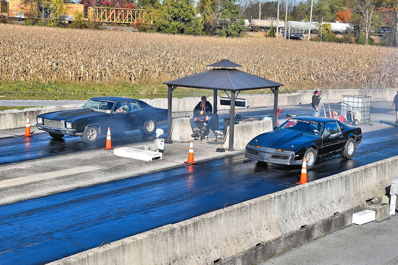 Two classic cars preparing to race on a drag strip, with bystanders and a shelter in the background, surrounded by a cornfield.