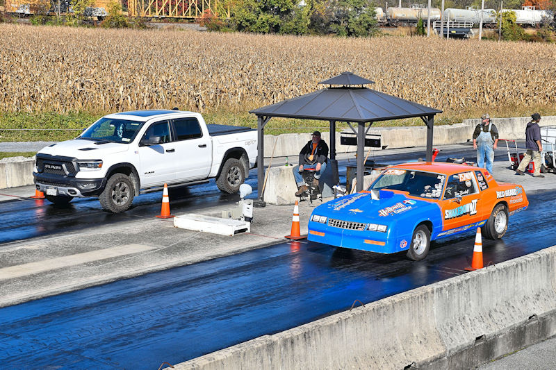 A white pickup truck parked next to a drag race starting line with an orange and blue racing car ready to race. Spectators are gathered under a gazebo nearby.