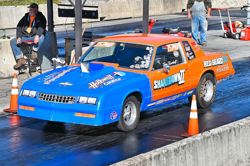 A brightly colored drag racing car in blue and orange, featuring sponsorship decals, positioned on a racetrack. In the background, a spectator is seated on a stool, while another person stands nearby. Traffic cones are placed around the car.