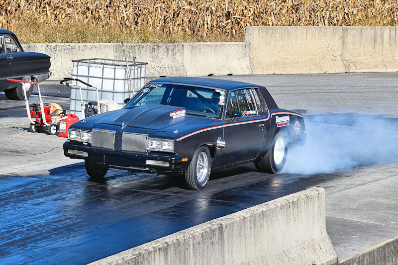 A black muscle car performing a burnout on a drag strip, with smoke billowing from its rear tires.