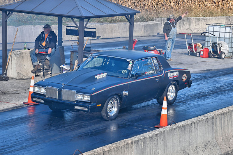 A black drag racing car lined up on a track with orange cones beside it, while a man sits in a chair watching and another man gestures beside a covered area.