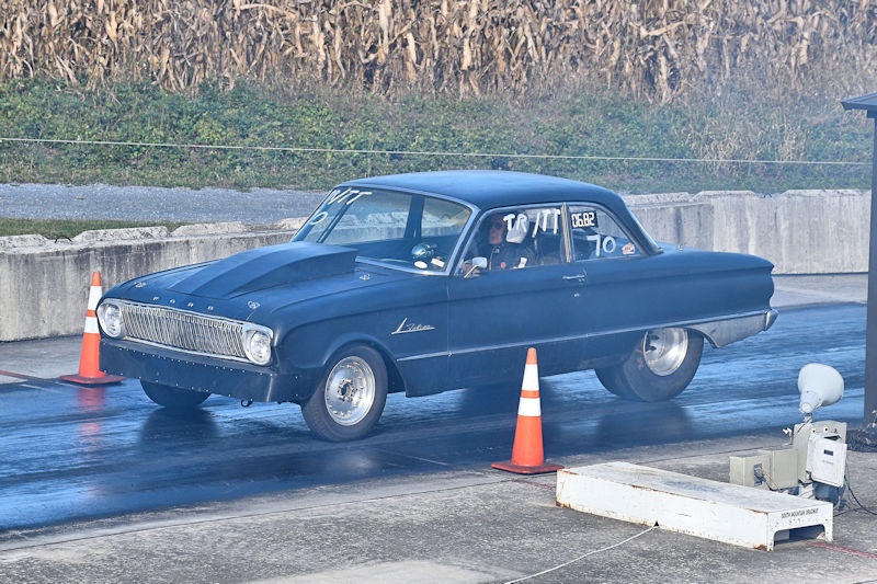 A vintage black car, possibly a 1960s model, is racing down a dragstrip, leaving smoke and tire marks on the asphalt. Traffic cones are positioned on the sides of the track.