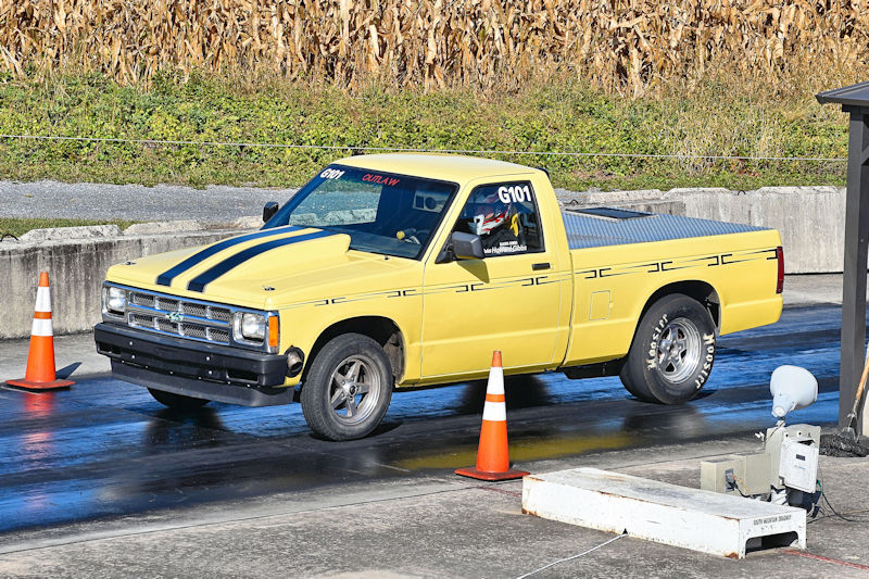 A yellow pickup truck with black racing stripes is driving down a drag strip, surrounded by orange traffic cones.