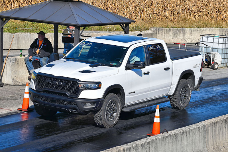 A white RAM pickup truck with off-road tires is lined up on a racetrack, with a man sitting under a gazebo nearby.