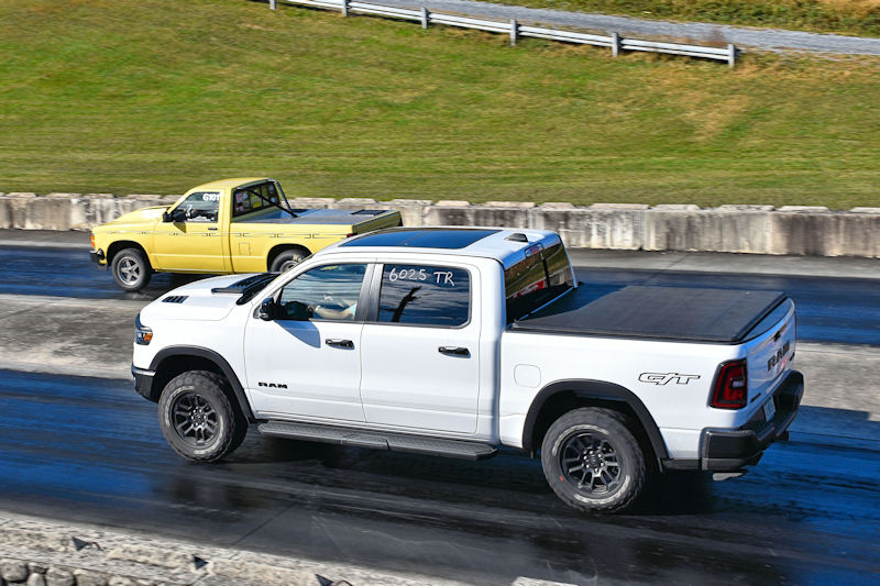 A white pickup truck racing alongside a yellow truck on a drag strip, with a green field and guardrails in the background.
