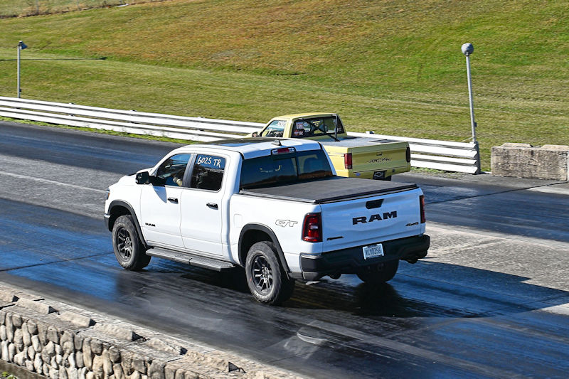 A white RAM pickup truck racing down a drag strip, with a yellow truck visible in the background.