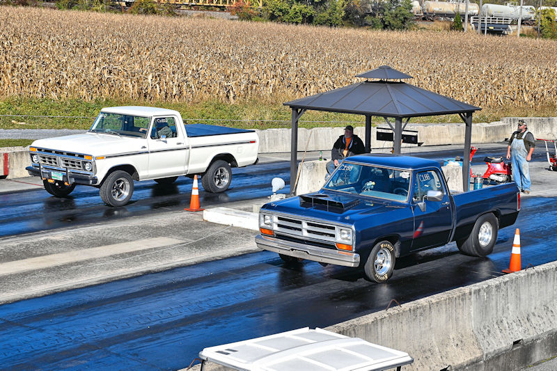 Two pickup trucks lined up for a drag race on a track, with a cornfield in the background and spectators nearby.