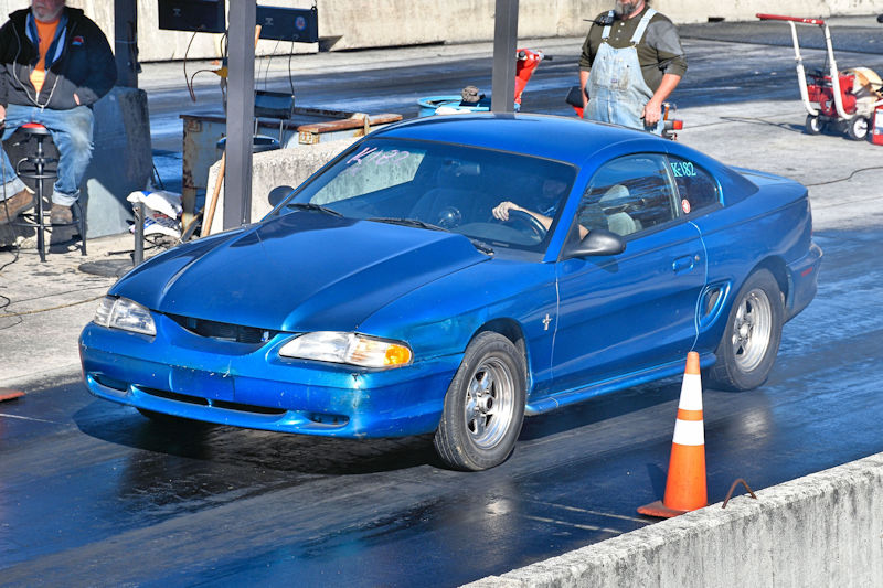 A blue car competing on a drag racing track, with a driver inside and a cone marking the track's edge. A person in the background watches from a stool.