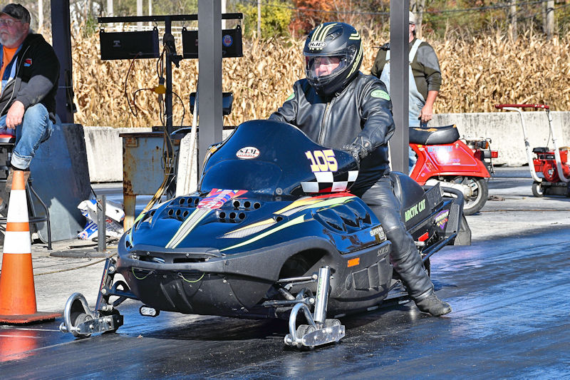 A racer in a black snowmobile with number 105 prepares for a drag race on a track, with a spectator sitting nearby.
