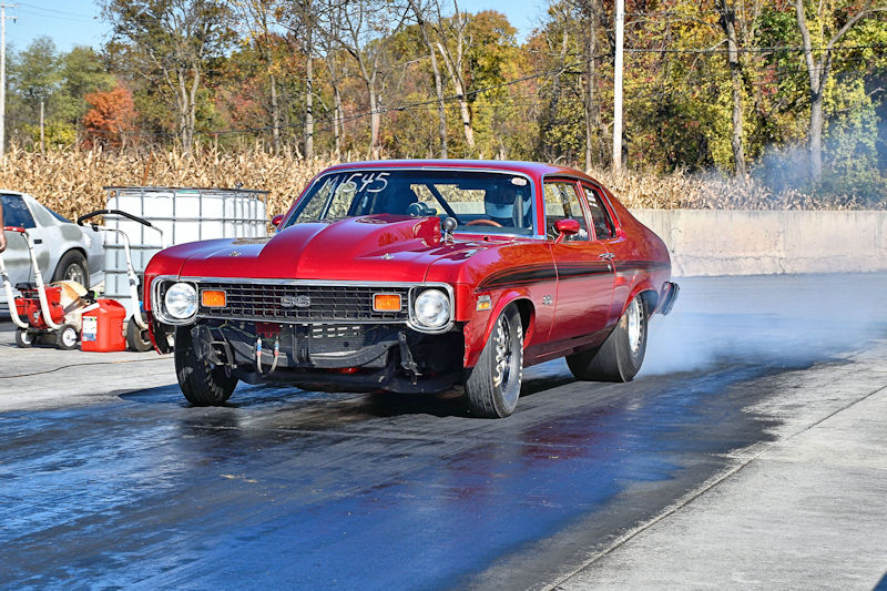 A red vintage muscle car racing on a drag strip, with tire smoke billowing from the rear tires and autumn foliage in the background.