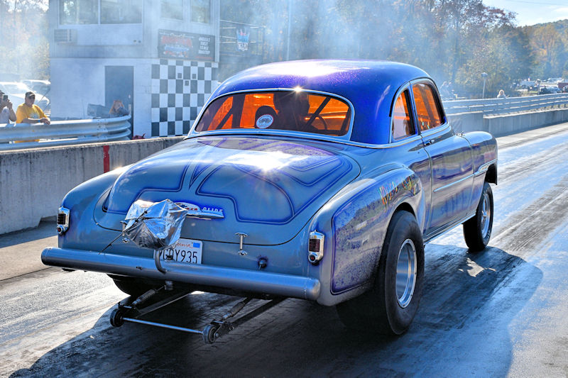 A vintage blue race car with a shiny finish is positioned on a drag strip, emitting smoke from the rear. The car features large rear tires and a chrome exhaust. In the background, spectators watch from the grandstands, and a checkered flag tower is visible.