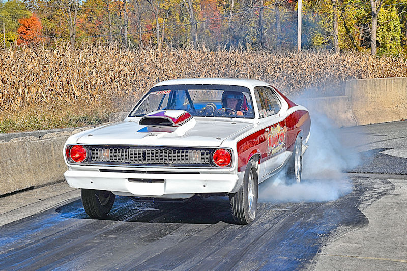 A classic drag racing car accelerates on a track, emitting smoke from the rear tires, with a backdrop of cornfields and autumn foliage.