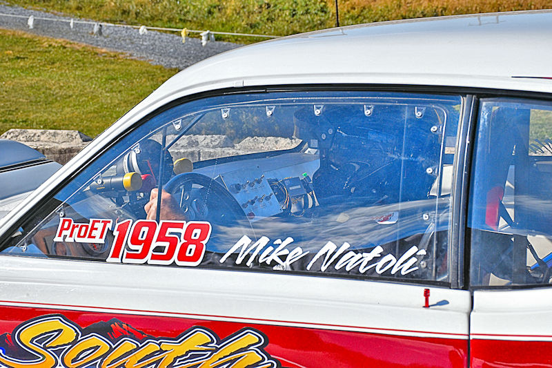 Close-up view of a race car's interior showing the driver holding the steering wheel, with dashboard controls visible and the driver's name and race number displayed on the side window.