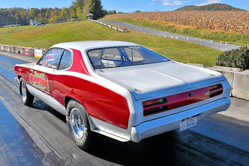 A classic muscle car with a red and white exterior parked on a drag strip, surrounded by greenery and a distant cornfield.