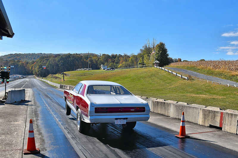 A classic white and red muscle car driving down a drag strip under a clear blue sky, with colorful autumn trees in the background.