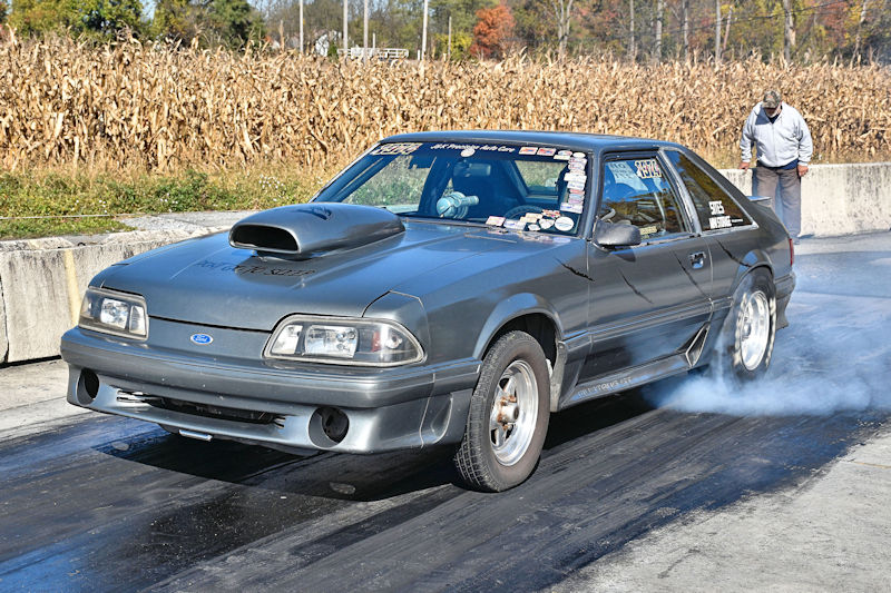A gray modified Ford Mustang racing on a drag strip, releasing smoke from the rear tires against a backdrop of cornfields.