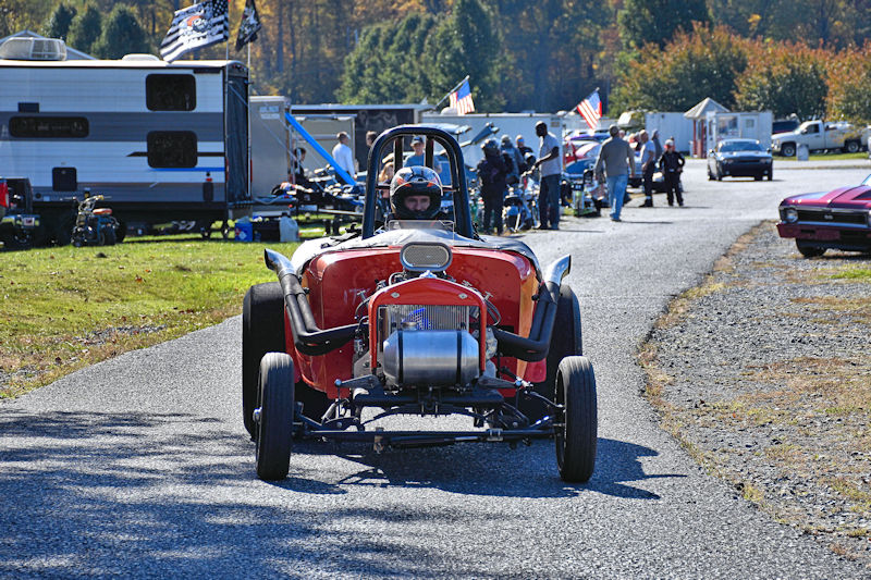 A vintage race car with an exposed engine drives on a gravel path, surrounded by people and parked vehicles at a racing event.