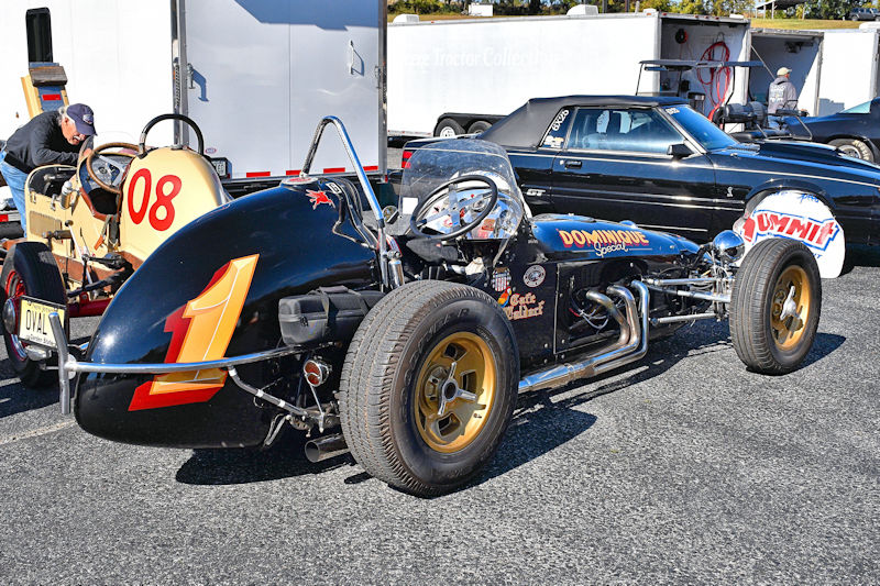 A vintage race car with a black body and prominent number '1' on the front, parked beside another small vehicle. The car features gold wheels and various decals. A man is crouching near the other vehicle in the background.