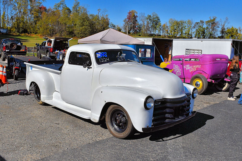 A vintage white pickup truck parked at an outdoor event, with a variety of other vehicles in the background, including a purple car with whimsical signage.