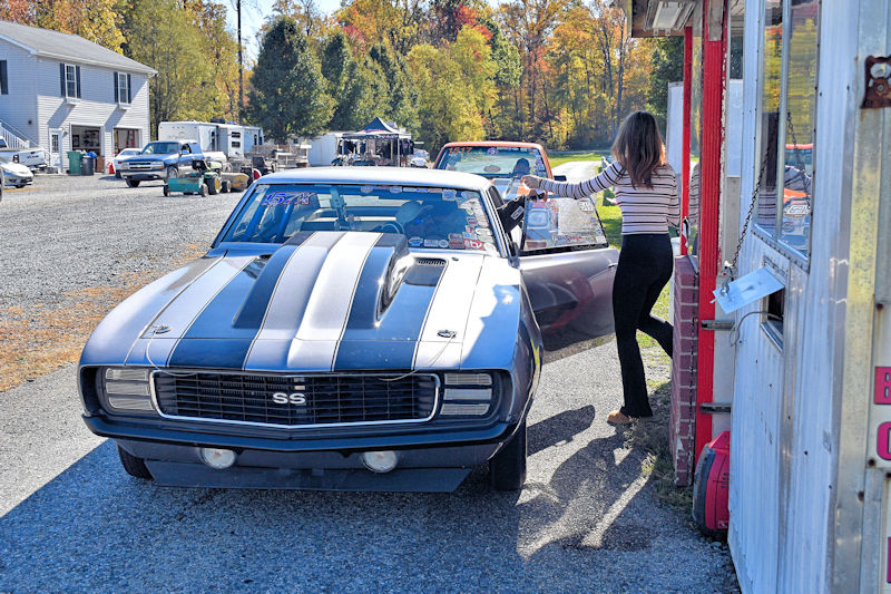 A vintage black and silver Camaro SS parked near a service area, with a woman in striped clothing opening the car door, set in a fall landscape.