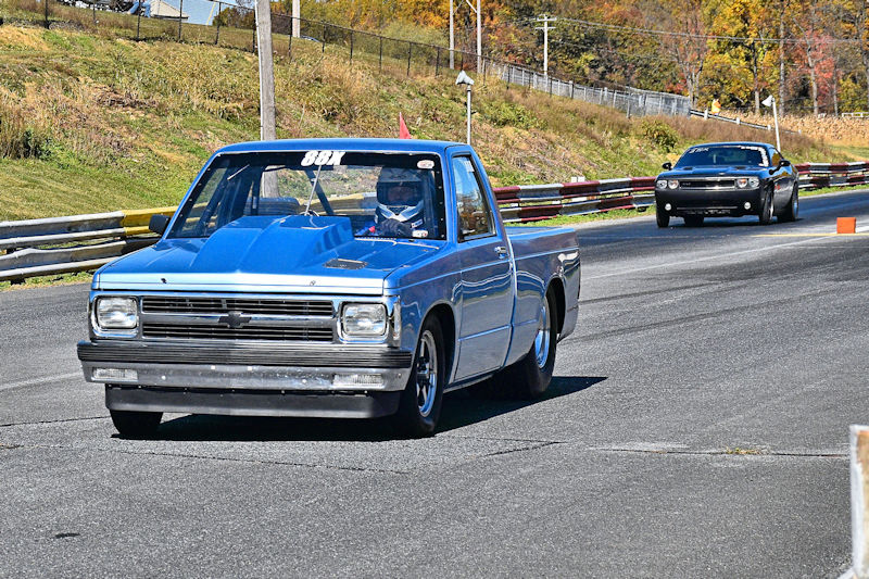 A blue custom pickup truck with a driver wearing a helmet races on a drag strip, with another car visible in the background.
