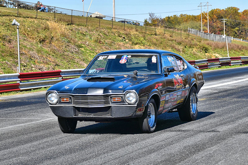 A black vintage car with racing stripes driving down a drag strip, showcasing its sleek design and competing stickers.