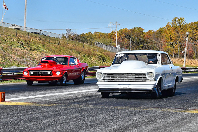 Two classic race cars, a red Mustang and a white sedan, racing on a drag strip.