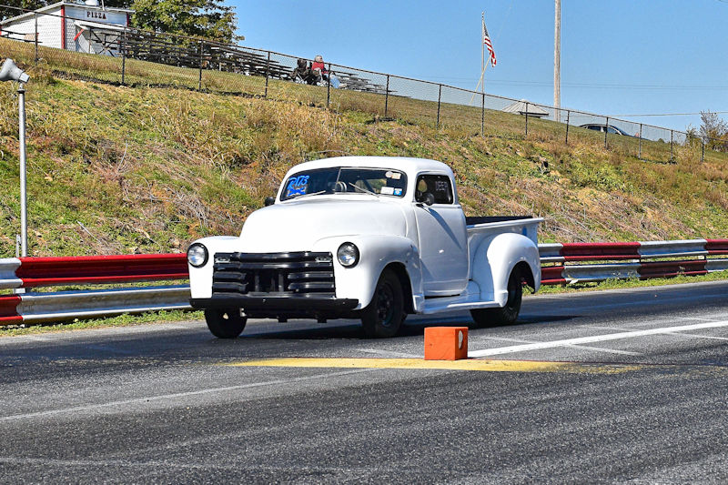 A vintage white pickup truck racing down a drag strip, with race timing lights visible in the foreground.
