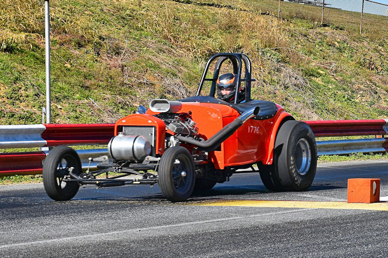 A classic drag racing car with an orange body, competing on a racetrack. The vehicle features an open design with a roll cage, and a driver wearing a helmet is seated inside.