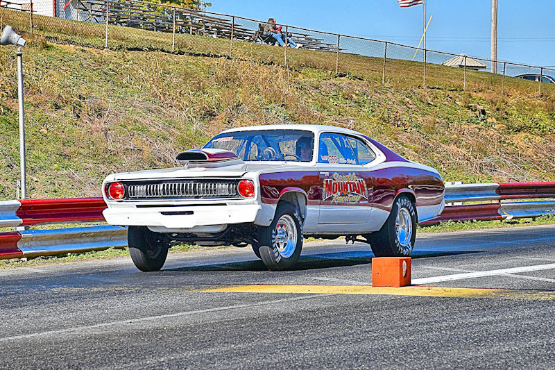 A classic muscle car launching off the starting line at a drag racing track, with its front wheels lifted off the ground and a crowd in the background.
