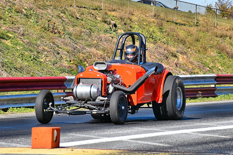 A vintage drag racing car in orange, driven by a helmeted racer, speeds down a track with a safety barrier alongside.