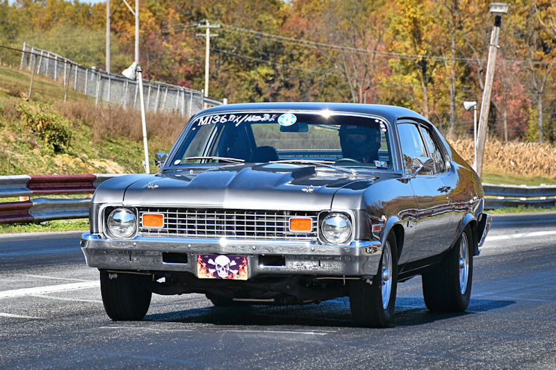 A shiny silver vintage car racing on a track, with autumn foliage in the background.