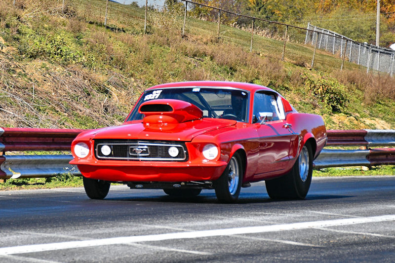 A red 1969 Ford Mustang racing down a drag strip with a visible front hood scoop and racing numbers on the side.