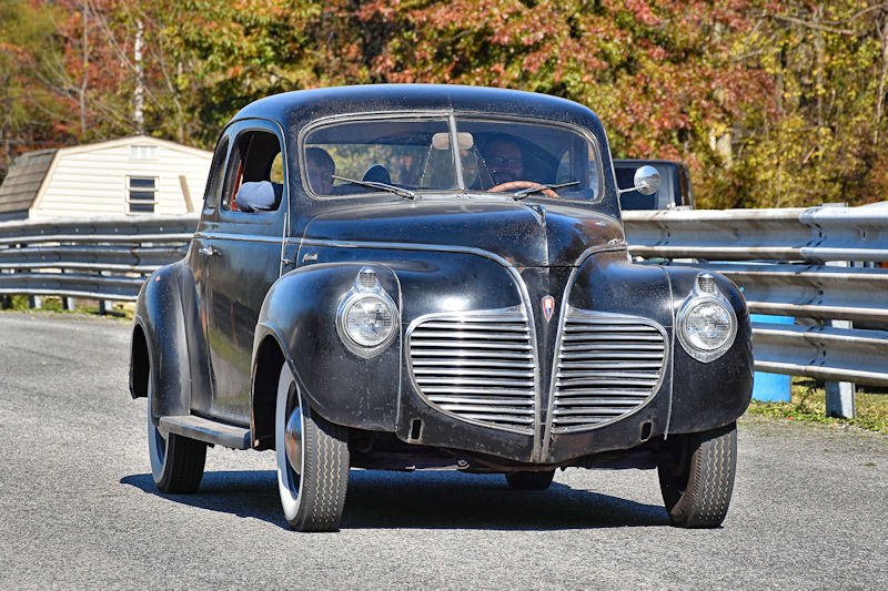 A vintage black car with a distinctive front grille driving on a racetrack surrounded by autumn foliage.