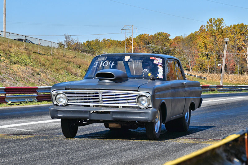 A vintage drag racing car with a matte finish, featuring a prominent hood scoop, driving down a racetrack with colorful fall foliage in the background.