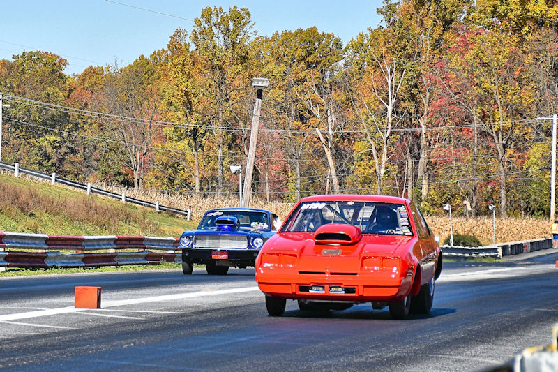 Two race cars competing on a drag strip, with vibrant autumn foliage in the background.