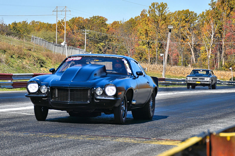 Two classic cars racing on a drag strip, with a black muscle car in the foreground and a silver car in the background, surrounded by fall foliage.