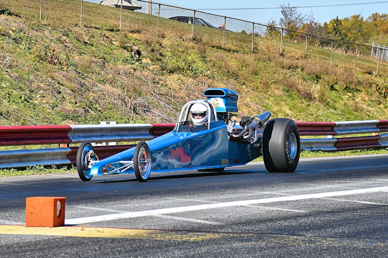 A blue dragster racing down a track, featuring a driver wearing a helmet. The scene includes safety barriers and grassy areas in the background.