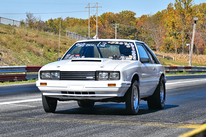A white racing car with multiple stickers on the windshield is driving down a drag strip surrounded by colorful autumn trees.